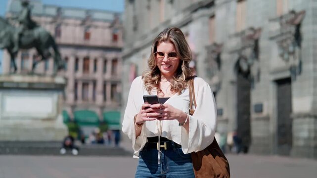 Young smiling Latina woman enjoys taking selfie outside Museo Nacional de Arte (MUNAL) in Mexico City, with the Equestrian Statue of Charles IV and neoclassical building in the background