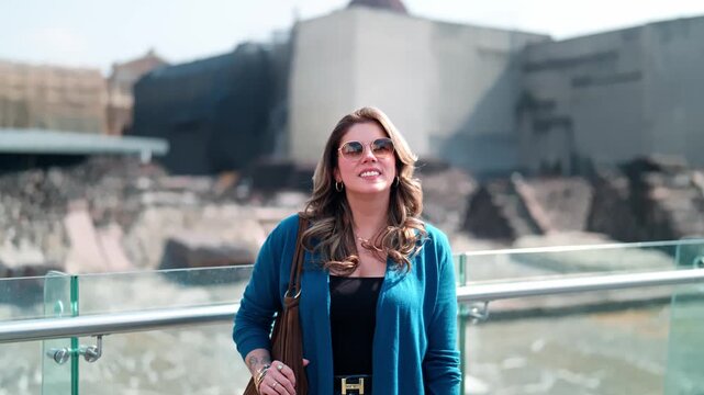 Young Latina woman smiles calmly next to the ancient ruins of Templo Mayor behind the Cathedral in Mexico City, wearing blue cardigan, jeans with bright sunlight and cultural landmarks surrounding her