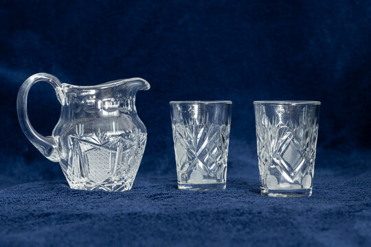 Cut crystal jug and two crystal shot glasses on dark blue fabric background. Clear engraved glass with geometric patterns. Studio still life photo.