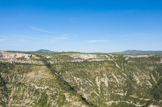 Vast limestone plateau with steep, green-speckled canyon walls under a clear blue sky at Cirque de Navacelles. Sunlight accentuates the rocky textures and natural contours of the landscape.