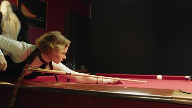 Young caucasian woman aims cue focused and strikes to a billiard ball on a red table. Dimly lit pool hall scene showing precise stance over red felt table. Poised shot and casual youth hangout
