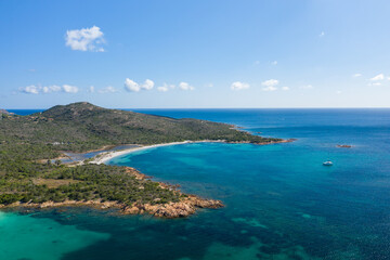 Obraz premium Aerial view of Spiaggia la Salinedda in Sardinia featuring vibrant turquoise waters, a white sandy beach, rocky shoreline, lush green hills, and a solitary yacht anchored offshore under a bright blue