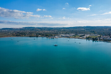 Obraz premium Aerial view of a peaceful marina and turquoise bay near Kilada, Greece. Calm water reflects scattered sailboats, with rolling hills and a partly cloudy sky in the background.