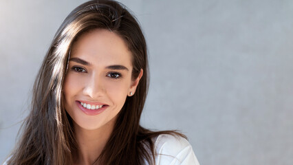 A woman with long hair smiles in a well-lit room. She stands in front of a light gray wall. The atmosphere is bright and inviting. Her expression shows joy and friendliness. © Prostock-studio