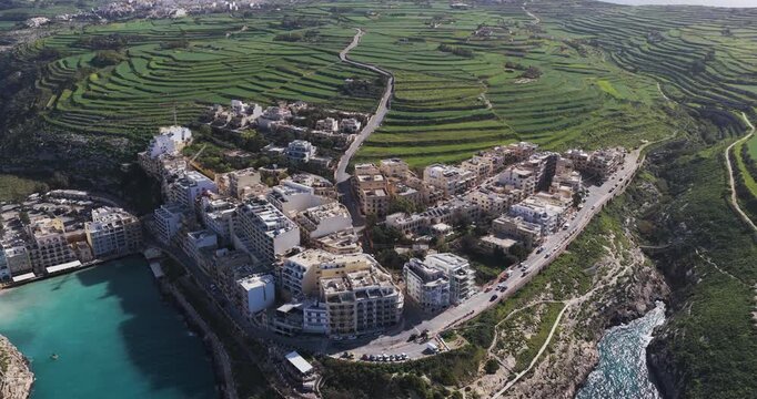 Aerial view of buildings clustered near the turquoise sea, contrasting with the terraced green landscape and rocky cliffs, Xlendi, Munxar, Malta.