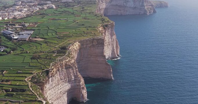 Aerial view of the rugged Xlendi cliffs meeting the deep blue ocean, contrasted by the bright green fields and the town in the background, Xlendi, Munxar, Malta.
