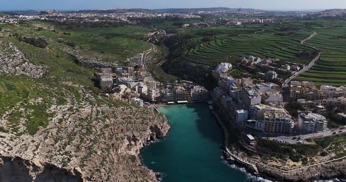 Aerial view of the turquoise waters meeting the rocky cliffs and buildings in Xlendi, creating a striking contrast against the green hills, Xlendi, Munxar, Malta.