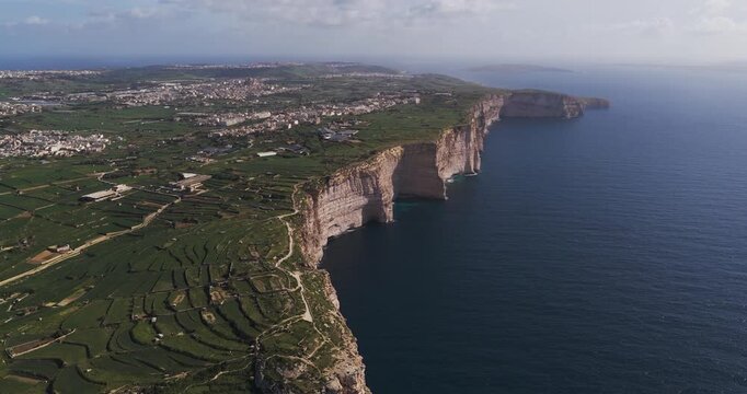 Aerial view of the dramatic Xlendi Cliffs plunging into the deep blue sea, contrasting against the lush green fields and distant cityscape, Xlendi, Munxar, Malta.