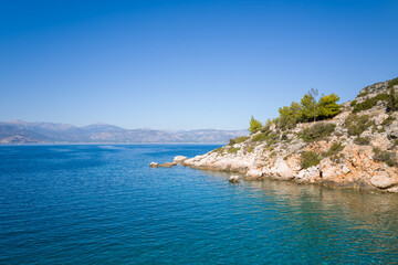Fototapeta premium Sunlit rocky headland covered with green pine trees extends into transparent blue water, with distant mountains under a vivid cloudless sky. The peaceful Mediterranean scene features textured rocks