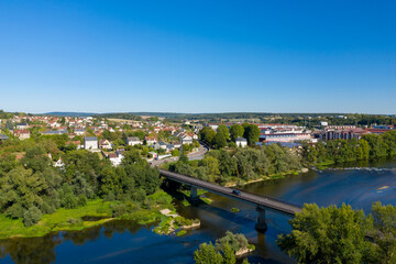 Fototapeta premium High-angle panorama of Imphy in central France, showing a bridge crossing the river, lush green trees, and residential rooftops under a vivid blue sky. The image highlights the tranquil rural