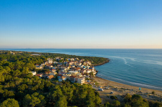 Aerial view of Arkoudi village with colorful houses nestled between green forest and sandy beach on the Ionian coast of Greece. Calm blue sea and clear sky create a peaceful Mediterranean atmosphere