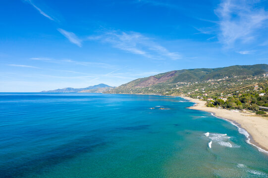 Expansive view of the vibrant blue Mediterranean Sea meeting a sandy beach and lush green hills near Palinuro, Italy. The scene is bathed in bright daylight with gentle waves and a tranquil, open