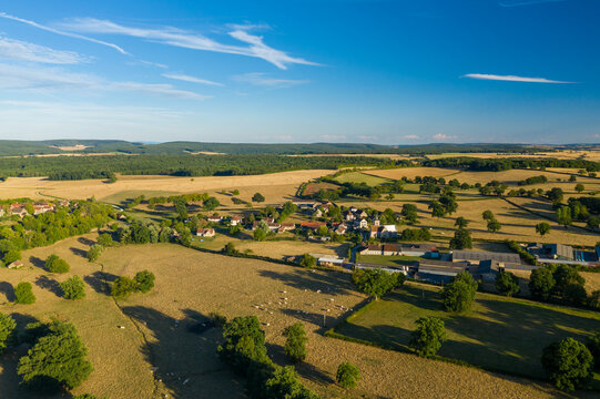Aerial view of Cuncy les Varzy village surrounded by sunlit golden fields, scattered trees, and rolling hills in the Burgundy region. The rural landscape features patchwork farmland, clusters of