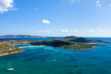 Fototapeta premium Drone perspective over Spiaggia Salina Bamba near San Teodoro, Sardinia, revealing vivid turquoise bays, anchored sailboats, and lush green hills under a clear blue sky.