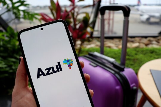 Bras&iacute;lia, Brazil - February 12, 2026: Person holding a smartphone with the Azul Linhas A&eacute;reas (Azul Brazilian Airlines) app with a suitcase in the background at the airport.