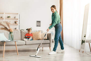Happy young mother mopping the floor and tidying up while her little son sitting on sofa and watching cartoon on smartphone, copy space