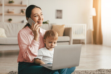 Happy mother with toddler son chatting on smartphone and using laptop computer, sitting on floor...