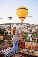 Fototapeta premium Woman stands on hotel rooftop observation deck with patterned pillows and woven rugs on terrace, watching hot air balloon fly over city at sunrise in Cappadocia, Turkey. Tourism and ballooning