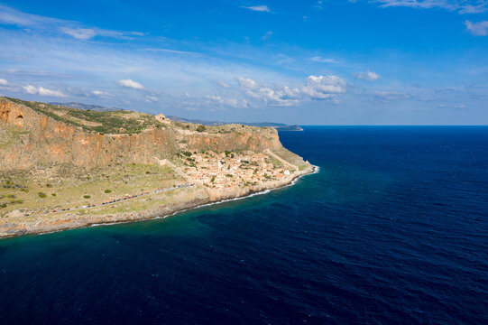 Aerial view of the historic Monemvasia village nestled beneath steep orange cliffs along the vivid blue Mediterranean Sea. The scene features rugged rock textures, clear water, and a bright sky with