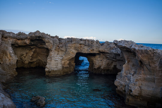 Natural stone arch formations rise above clear turquoise water along the rugged coastline of Marina Serra, Italy. The scene features textured limestone cliffs and a tranquil blue sea under an open sky