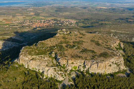 Aerial view of a rugged limestone plateau surrounded by sparse vegetation, overlooking a traditional village and distant coastline under clear afternoon light near Opoul Perillos.