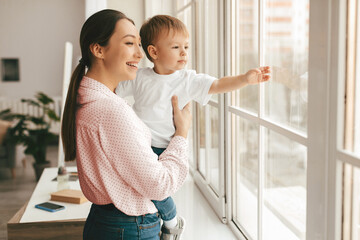 Happy young mother holding toddler son on hands, standing by window showing him world outside, kid...