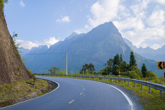 Curved mountain road bordered by a steep rocky slope and lush greenery, with dramatic peaks rising under a bright blue sky. Sunlight highlights the rugged landscape and creates a sense of open journey
