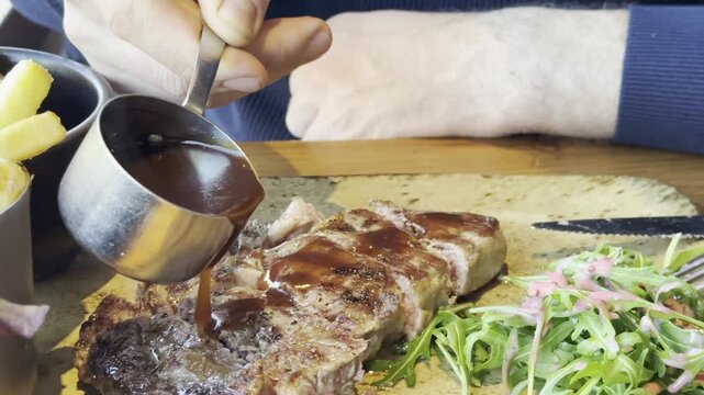A Caucasian man pouring sauce over a steak. Close-up of steak, salad, and French fries. Food sauce. Lunch at a cafe.