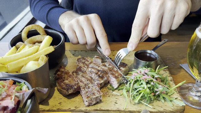 A white man cutting a steak. Close-up of a cafe table with food. Steak, fries, onion rings, and salad for lunch. Mealtime.