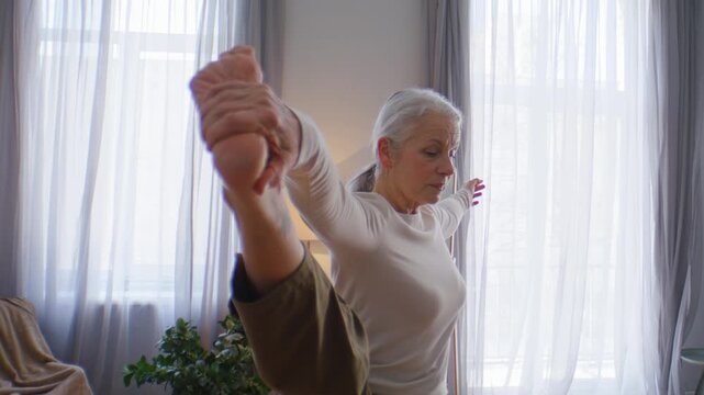 Senior woman grips raised bare foot holding strong balance pose