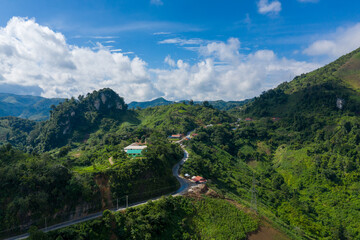 Fototapeta premium Curving mountain road winds past scattered houses and dense green hills under a vivid blue sky with scattered clouds near Phou Khoun, Laos. The scene features dramatic terrain, vibrant vegetation, and