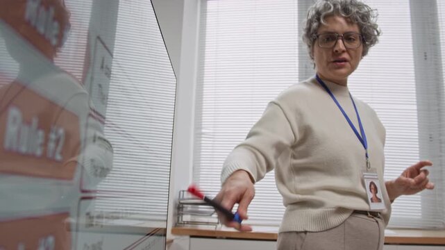 Close up shot of grey haired mature woman as female teacher explaining English grammar rules during class with focus on hand pointing at digital screen, copy space