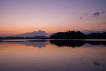 Fototapeta premium Soft morning light illuminates calm waters at Lac des Settons, with gentle pastel colors in the sky and a dark forest silhouette mirrored on the tranquil lake surface.