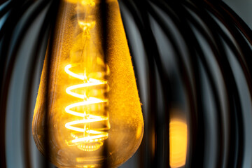 Spiral warm filament of a glowing bright light bulb inside a lampshade. The illuminated coil creates a cozy and inviting atmosphere with soft amber light.