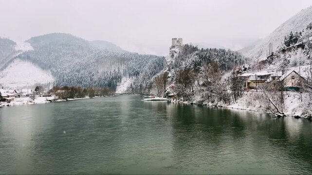 Medieval Strecno Castle on a snowy cliff above the Vah river in winter. Historic 4K landscape of a Slovakian fortress with an old river ferry and mountains in the Mala Fatra region, Slovakia.