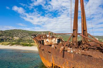 Fototapeta premium The rusted bow of the Dimitrios shipwreck rises above shallow clear water with sandy beach and green hills in the background near Gythio, Greece. Morning light highlights the corroded textures and