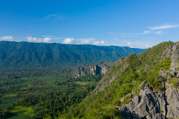 Fototapeta premium Expansive view of a rugged limestone ridge covered in lush vegetation, overlooking a wide green valley and distant forested mountains under a clear blue sky. The scene features sharp rock textures
