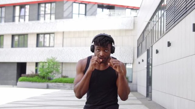 African American man in sportswear boxing outdoors with headphones, athletic guy practicing punches and doing shadow boxing on urban background
