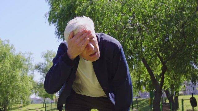 tired elderly man is exercising in the park and is starting to catch his breath, exhausted gray-haired pensioner is suffering from the heat after running outdoors