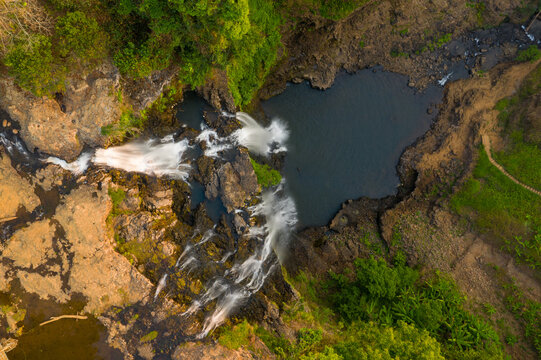 Top-down view reveals Tad Yuang waterfall cascading over rugged basalt rocks into a dark pool, surrounded by lush green vegetation on the Bolaven Plateau in southern Laos. Warm sunlight enhances the