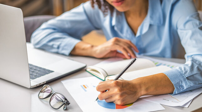 Woman analyzing financial report and writing notes on paper charts beside laptop. Business planning, data analysis, strategy, accounting, productivity, home office
