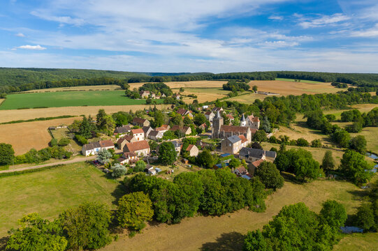 Sweeping aerial perspective of Beuvron, a rural village surrounded by patchwork fields, rolling hills, and dense woodland. The historic stone church and traditional houses nestle amid lush summer