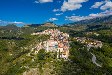 Obraz premium Aerial perspective of Rivello, Italy, reveals a historic hilltop village with terracotta roofs cascading down a lush green slope, surrounded by dramatic mountains under a vibrant blue sky. Crisp