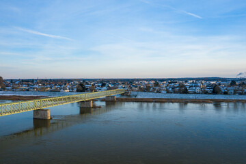 Fototapeta premium A long pale green iron bridge stretches across the calm Loire River towards a village dusted with snow, under a soft blue winter sky in Sully sur Loire.