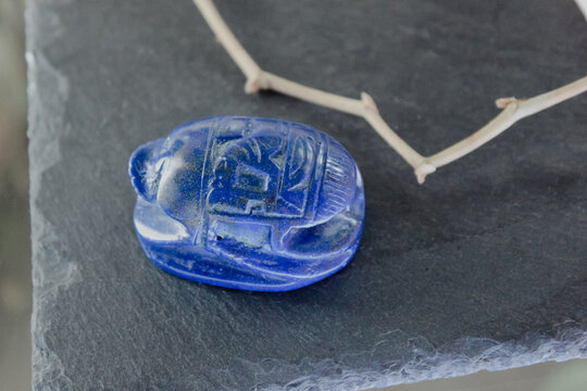 Lapis lazuli scarab beetle carving on dark slate stone with decorative dry branches, ancient amulet.