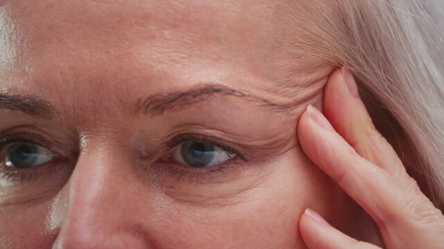 Close up: old woman eye. Middle aged female wrinkles near eyes. Aging, old skin, wrinkled face beauty portrait. Elderly lady touches skin on temples with fingers, facelift cosmetic cream. dermatology