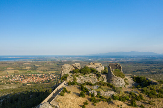 Weathered stone walls of a ruined fortress stand atop a sunlit hill, overlooking a vast plain, distant coastline, and a small village under a clear blue sky near Opoul Perillos.