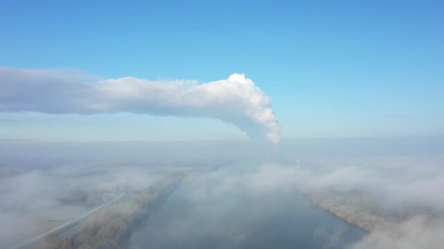 Drone shot of steam rising from the Dampierre nuclear power plant in France, with a river and foggy landscape under a clear blue sky.
