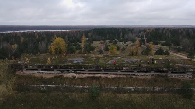 Aerial drone view shows Dretunsky military range bunker, people walking, grassy berms, sparse pine and birch, dirt track, white van, lake, overcast skies.