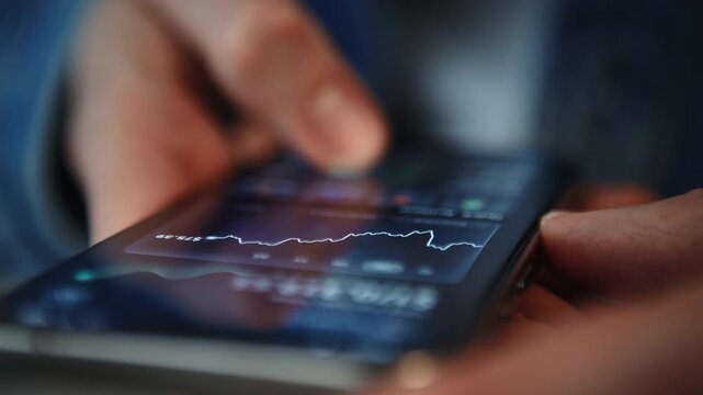 Close-up of male hand in blue denim shirt holding smartphone with financial app showing portfolio growth on screen. Indoor daylight scene. Investing, mobile finance, wealth growth, fintech technology